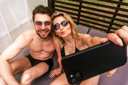 Photo for Blonde lady taking selfies with her happy companion while sitting on the mattress in a gazebo. Leisure activity and summer vacation concept - Royalty Free Image
