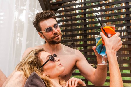 Photo for Blonde young woman and her boyfriend clinking glasses with cocktails while resting in a gazebo. Romance and summer vacation concept - Royalty Free Image
