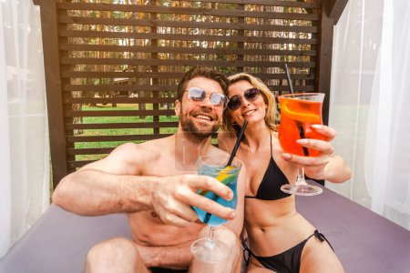 Photo for Cheerful young couple of vacationers in sunglasses seated in the pergola with a glasses of cocktails posing for the camera - Royalty Free Image