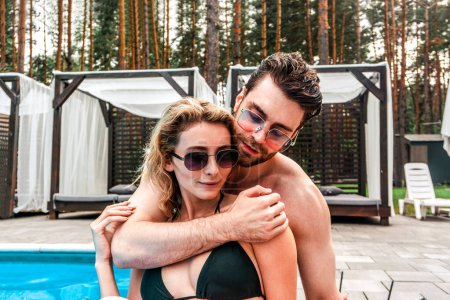 Téléchargez les photos : Beau jeune homme amoureux passionné embrassant son compagnon féminin attrayant près d'une piscine extérieure. Romance et vacances d'été concept - en image libre de droit