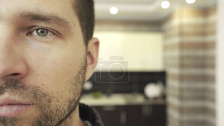 Young bearded man looking at camera, standing at kitchen at home. Half face. Copy space. Emotion, despair concept. Slow motion
