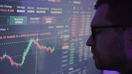 Businessman trader in glasses looking at big screen with trading charts. Slow motion. Stock market, trade, investment, technology concept. Close up