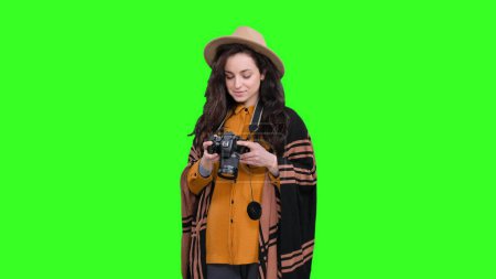 Junge Frau mit Hut posiert mit Kamera und winkt mit der Hand auf grünem Hintergrund. Reise, Tourismus, Abenteuerkonzept