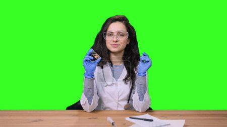 Female doctor in uniform with stethoscope in protective gloves in laboratory holding vaccine bottle on hand on green isolated background. Health medical concept
