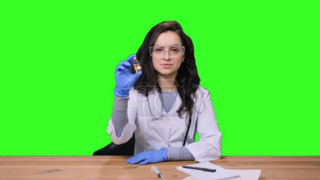 Female doctor in uniform with stethoscope in protective glasses holding vaccine bottle, sitting at desk on green isolated background. Health care, medicine concept