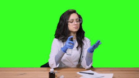 Young female doctor in uniform and protective glasses disinfecting hands, sitting at desk on green isolated background. Health care, medicine concept