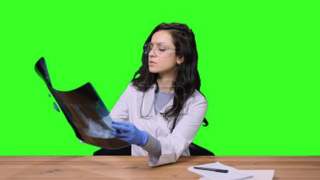 Female doctor wearing white medical gown suit, working in hospital clinic office, holding X-ray on green isolated background. Health medical concept