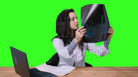 Female doctor in white medical uniform, working with laptop in clinic office, holding at X-ray on green isolated background. Health care, medicine concept