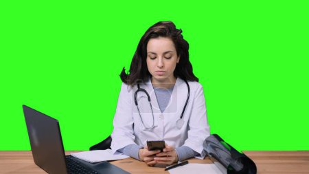 Female doctor in uniform typing on the mobile phone while working at desk with laptop on green isolated background. Health care, medicine concept