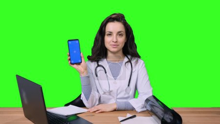 Happy young female doctor working with laptop while holding mobile phone, screen for copy space on green isolated background. Health care, medicine concept