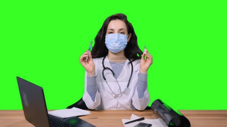 Female doctor in protective face mask shoeing vaccine and syringe while sitting at desk in office on green isolated background. Health care, medicine concept