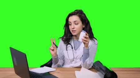Female doctor holding bottle with medicine while talking online with patient by the laptop on green isolated background. Health care, medicine concept