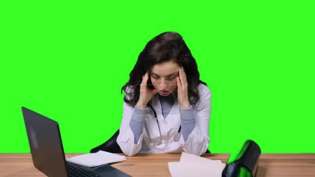 Tired young female doctor having headache while wortking at office, sitting at desk, using laptop on green isolated background. Health care, medicine concept