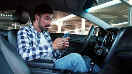 Feliz joven leyendo el mensaje en el teléfono inteligente mientras está sentado en el coche. Transporte, tecnología, estilo de vida