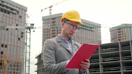 Waist-up portrait of site supervisor writing on clipboard while standing near tower cranes and unfinished multi-story houses. Slow motion