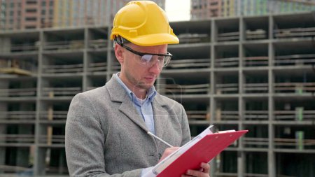 Waist-up portrait of focused construction auditor making notes in documents on clipboard near unfinished multi-story house. Slow motion