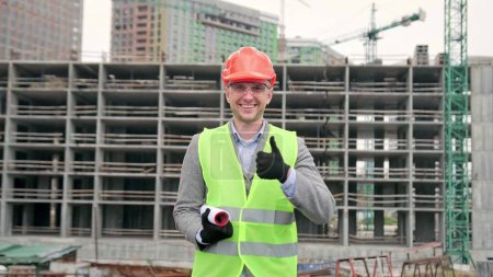 Project manager with drawings giving thumbs-up and smiling at camera while standing near unfinished multi-story house. Dolly shot