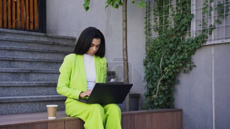 Señora americana sentada afuera, sosteniendo el portátil y escribiendo, trabajando en línea y bebiendo café. En cámara lenta. Concepto empresarial