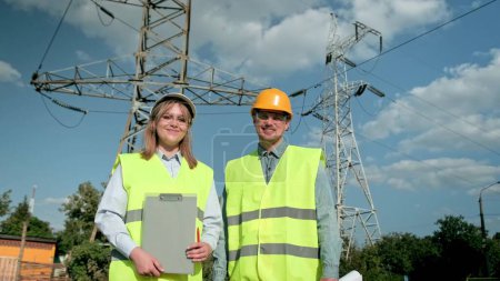 Smiling power line inspectors with clipboard and pleased colleague with drawings standing near transmission towers during site inspection. Dolly shot