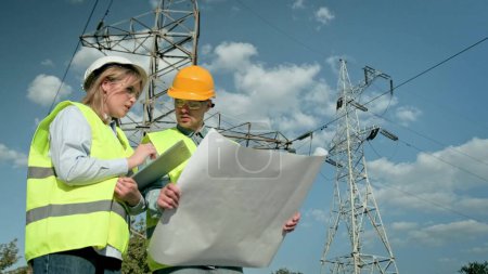 Power engineer making notes on clipboard and showing something to coworker with blueprints while standing near transmission towers. Slow motion
