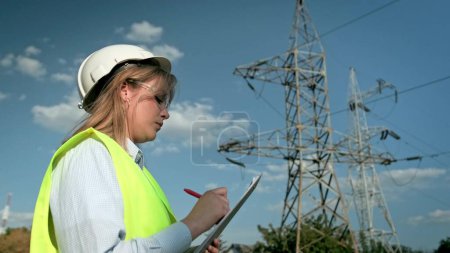 Serious energy auditor in hard hat making notes on clipboard while pointing at transmission tower during on-site audit. Slow motion