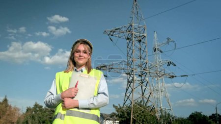 Waist-up portrait of joyous energy auditor in hard hat with clipboard and pen standing near electrical transmission towers. Dolly shot