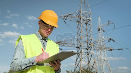 Waist-up portrait of energy auditor in hard hat writing something on clipboard while standing near electricity pylons. Slow motion