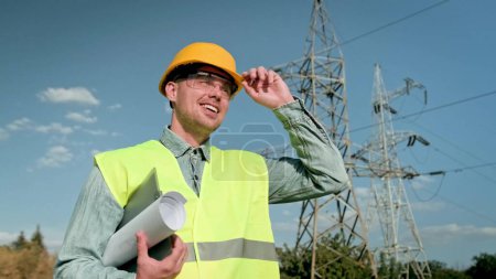 Waist-up portrait of cheerful power line inspector with blueprints touching his hard hat while walking past transmission towers. Slow motion