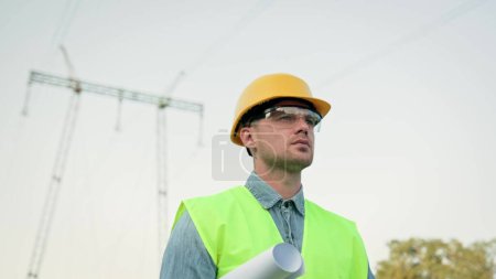 Portrait of serious energy auditor with drawings under arm moving away from transmission tower after on-site audit. Slow motion