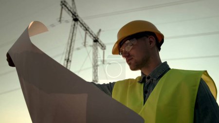 Portrait of power line inspector in hard hat and goggles looking at drawings while inspecting electricity pylon outdoors. Slow motion