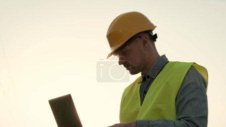 Portrait of energy auditor working on portable computer while standing near electricity pylon. Slow motion