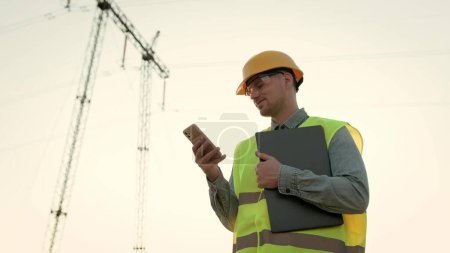 Waist-up portrait of pleased energy auditor with laptop reading something on smartphone while standing near electricity pylon. Slow motion
