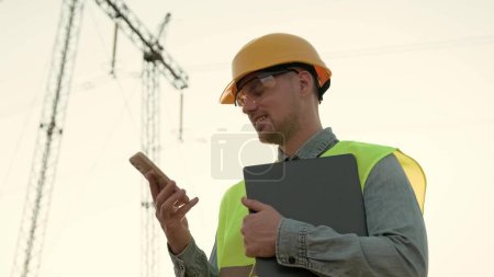 Portrait of power line inspector holding laptop and smiling while talking on cellphone near transmission tower. Slow motion