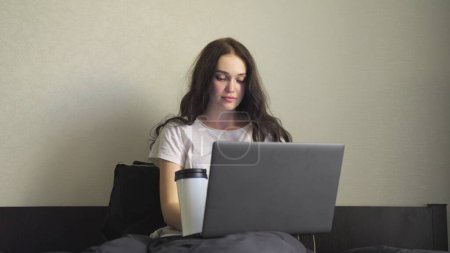 Junge Frau arbeitet online am Laptop, trinkt Kaffee und sitzt im Bett. Zeitlupe. Arbeiten zu Hause Konzept. Innenbehörde