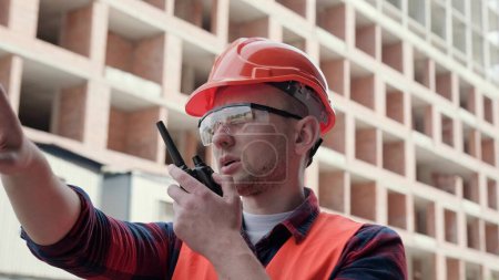 Der Bauleiter steht in der Nähe eines unfertigen mehrstöckigen Gebäudes, zeigt mit dem Finger auf etwas in der Ferne und redet auf einem Walkie-Talkie. Zeitlupe