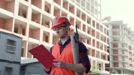 Serious construction superintendent writing something on clipboard while looking at unfinished multi-story houses. Slow motion