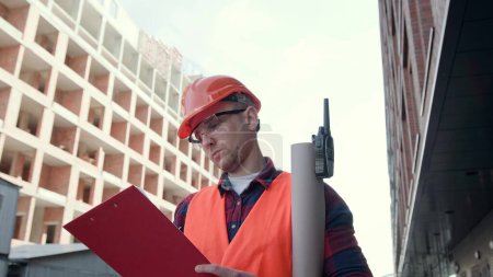 Waist-up portrait of focused foreman writing something on clipboard while standing between unfinished multi-story houses. Dolly shot