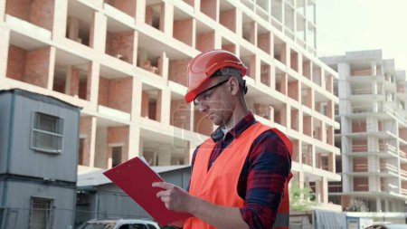 Waist-up portrait of focused construction supervisor writing something on clipboard while standing between unfinished multi-story houses. Slow motion