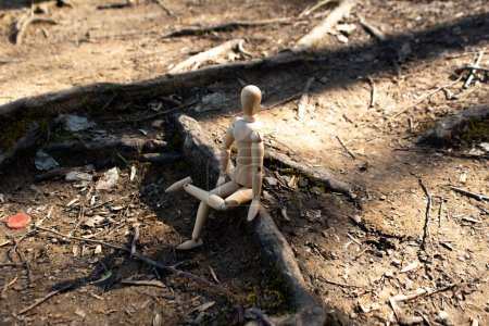 Téléchargez les photos : Mannequin en bois assis sur des racines d'arbre regardant vers la forêt, jambes de marionnette en bois croisées, vue latérale, concept d'automne, arrière-plan de la forêt - en image libre de droit