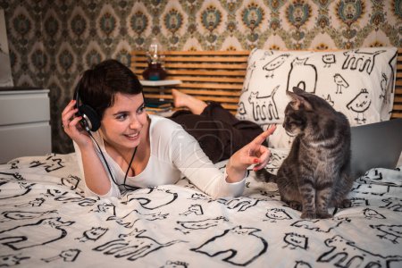 Photo for Beautiful woman plays with the cat on the bed. The young woman listens to music on headphones while she is petting her cat. - Royalty Free Image