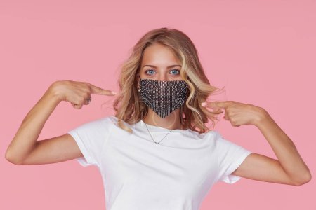 Foto de A young attractive woman in a white casual t-shirt wears  a stylish fashionable protective face mask. Protection against flu and cold diseases during a pandemic. Pink isolated background - Imagen libre de derechos