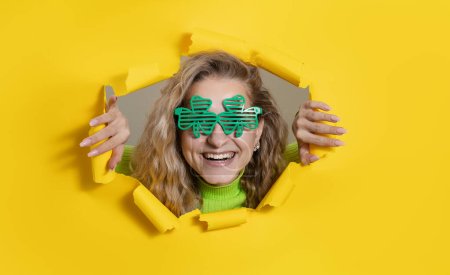 Foto de Mujer sorprendida con gafas verdes en forma de trébol mirando, mirando a través del agujero de papel amarillo brillante. Mostrando mano a lado. Copia espacio para texto. Día de San Patricio. Wow cara - Imagen libre de derechos