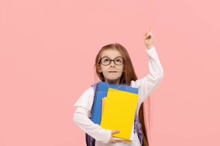 Photo for Portrait of a schoolgirl with textbooks and a backpack on a pink background points up. Back to school - Royalty Free Image
