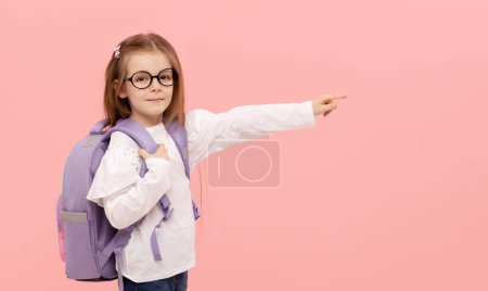 Photo for Portrait of a schoolgirl with textbooks and a backpack on a pink background pointing to the right. Back to school - Royalty Free Image