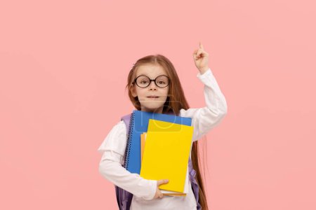 Photo for Portrait of a schoolgirl with textbooks and a backpack on a pink background points up. Back to school - Royalty Free Image