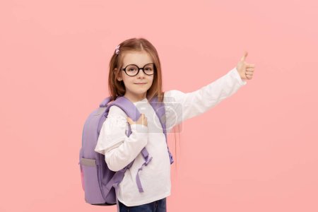 Photo for Portrait of a schoolgirl with textbooks and a backpack on a pink background pointing to the right. Back to school - Royalty Free Image