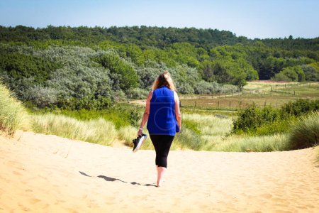 Téléchargez les photos : Un superbe paysage d'une femme marchant sur les dunes de sable de la plage vers la forêt. Le temps est très chaud, comme tel la femme a enlevé ses chaussures et les porte. - en image libre de droit