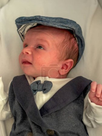 Un hermoso retrato de bebé de un recién nacido. El niño está vestido con elegancia, con un traje de tres piezas, corbata de lazo y gorra plana.
