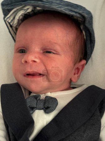 Ein wunderschönes Babyporträt eines neugeborenen Jungen. Das Kind ist elegant gekleidet, trägt einen dreiteiligen Anzug, Fliege und Schiebermütze.