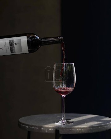 Foto de Persona vertiendo vino tinto de la botella en una copa de vino sobre una mesa de hormigón, fondo oscuro - Imagen libre de derechos